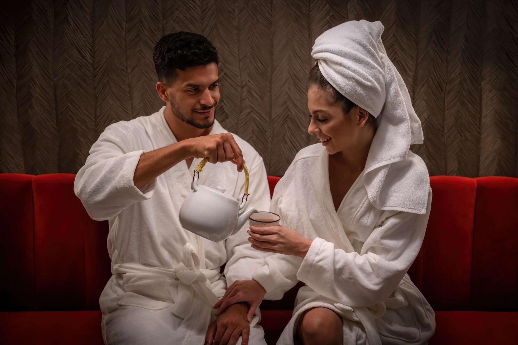 Radisson Blu Hotel, Dubai Deira Creek Exterior photo The photo depicts a man and a woman sitting together on a red couch, both dressed in white bathrobes. The man is pouring tea from a white teapot into a cup held by the woman, who is smiling. She has a towel wrapped around her head, suggesting a relax