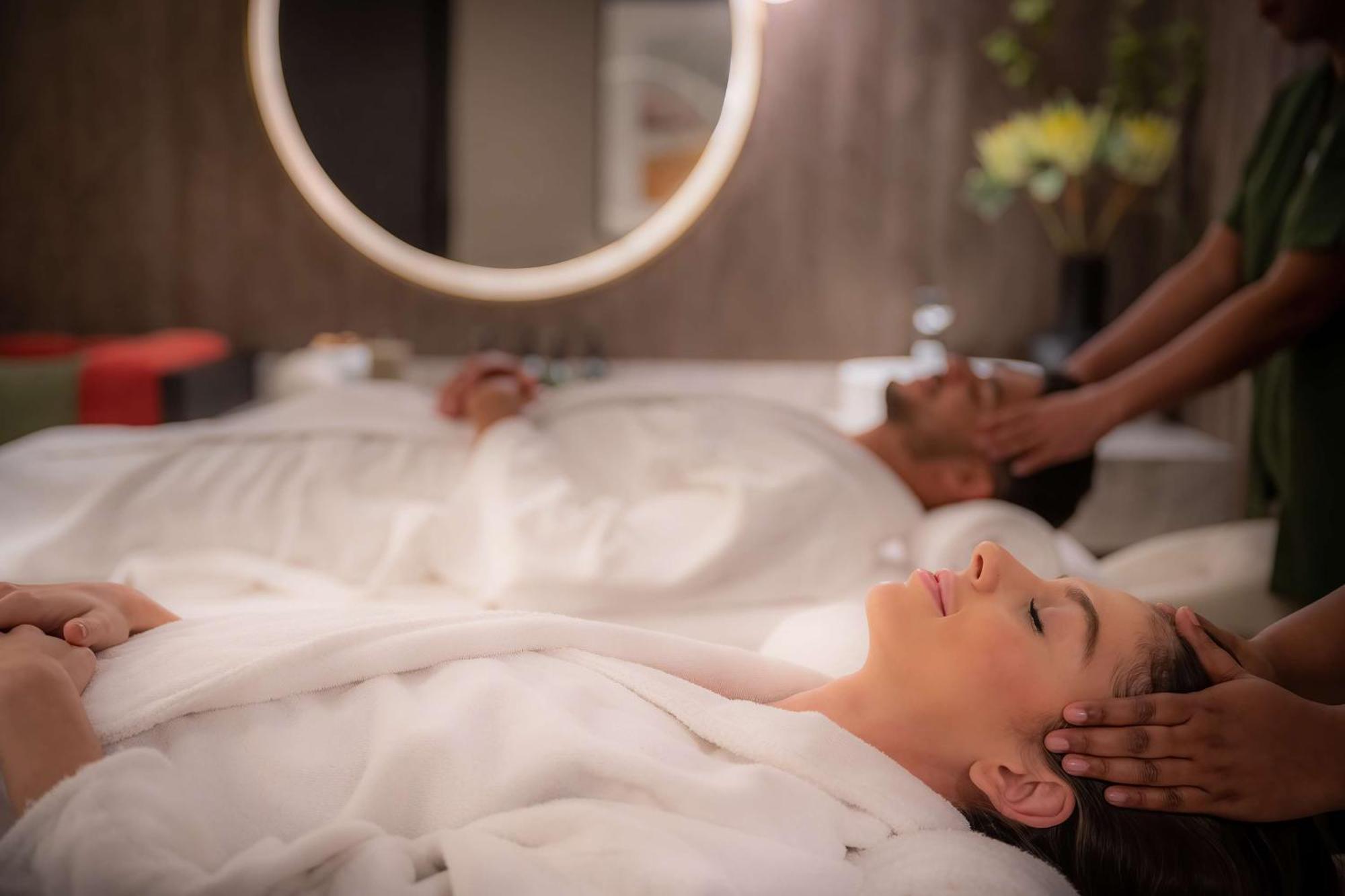 Radisson Blu Hotel, Dubai Deira Creek Exterior photo The photo shows a serene spa setting where a woman and a man are lying on treatment tables. Both are wearing white robes and appear to be undergoing a relaxing treatment. The woman has her eyes closed and seems to be enjoying a facial massage, while 