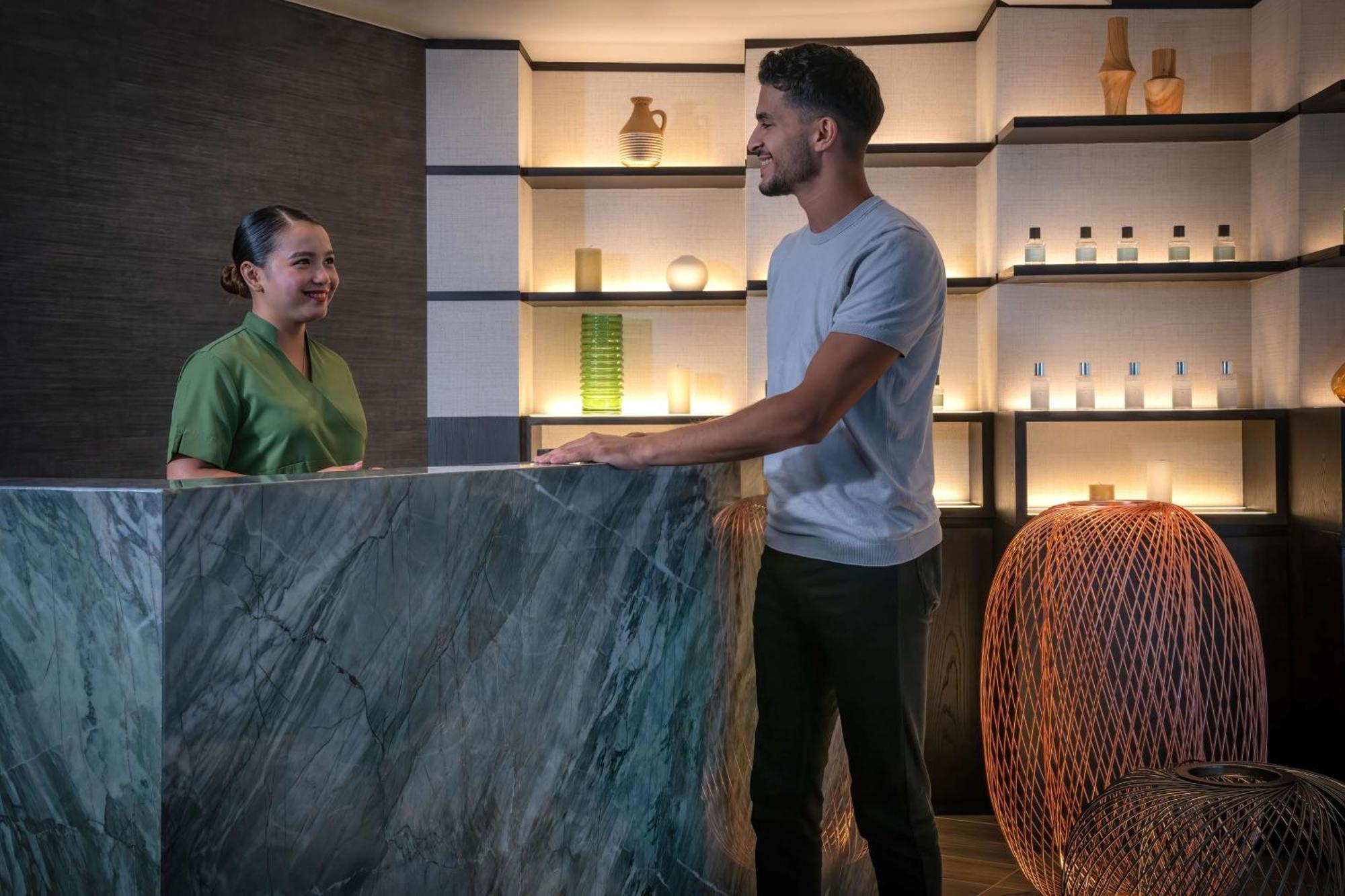 Radisson Blu Hotel, Dubai Deira Creek Exterior photo The image shows a reception area, likely in a spa or wellness center. On one side, there is a woman wearing a green uniform, smiling at a man who is standing at a marble reception desk. The man is dressed casually in a gray t-shirt and dark pants. Be